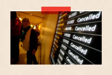 Getty Images Wide-angle view inside an airport terminal showing a large electronic departures board on the right with multiple flight listings, each marked ‘Cancelled’ in bright white text against a dark background, with flight numbers visible. To the left, several travelers walk past the display along a softly lit corridor, with luggage and coats partially visible. The perspective emphasizes the repeated cancellation notices.