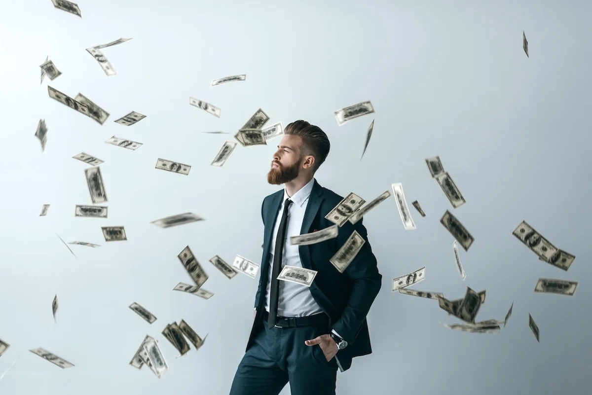 Best Savings Accounts Daily Bearded man in a dark business suit standing confidently with his hand in his pocket while US dollar bills rain down around him against a gray background. This illustrates the potential for wealth accumulation and passive income through high-yield savings accounts, which are currently offering interest rates up to 5.00% APY as of June 2026, allowing savers to maximize returns despite fluctuating market conditions. Source: The College Investor