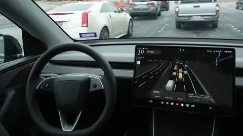 Getty Images A black steering wheel and large screen to the right which shows lanes of traffic and vehicles in it.