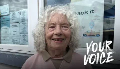 BBC Close-up of Maggie Dodd with curly grey hair wearing a pink top, standing in front of a post office window with notices and a “Track it” Royal Mail sign visible in the background.
