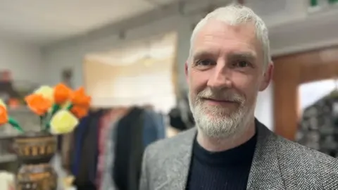 Close-up of Scott McBride with short grey hair and a trimmed beard wearing a textured blazer and dark top, standing indoors in front of a clothing rack and shelves, with a vase of orange and cream flowers visible to the side.