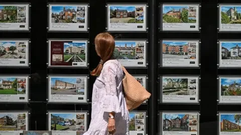 Getty Images A woman in a white flowy dress walks past an estate agent window filled with home listings.