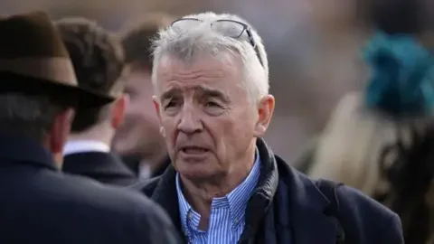 PA Media Michael O'Leary, a man with grey hair, wearing a blue jacket and a blue and white striped shirt. He has glasses on top of his head.
